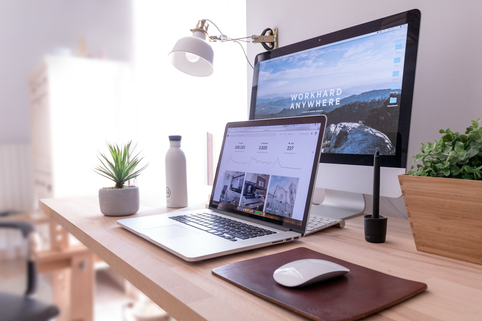 A laptop and desktop monitor on a bright wooden desk beside plants.